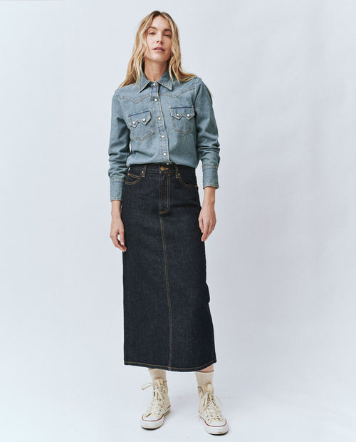 Woman wearing a light blue button up denim shirt and a dark blue long denim skirt.