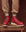 A person wearing vibrant Chili Pepper Red Wing Heritage Boots from the THE GREAT. HOL 24 RED WING collection and beige pants stands confidently on a wooden stool against a rich brown background.