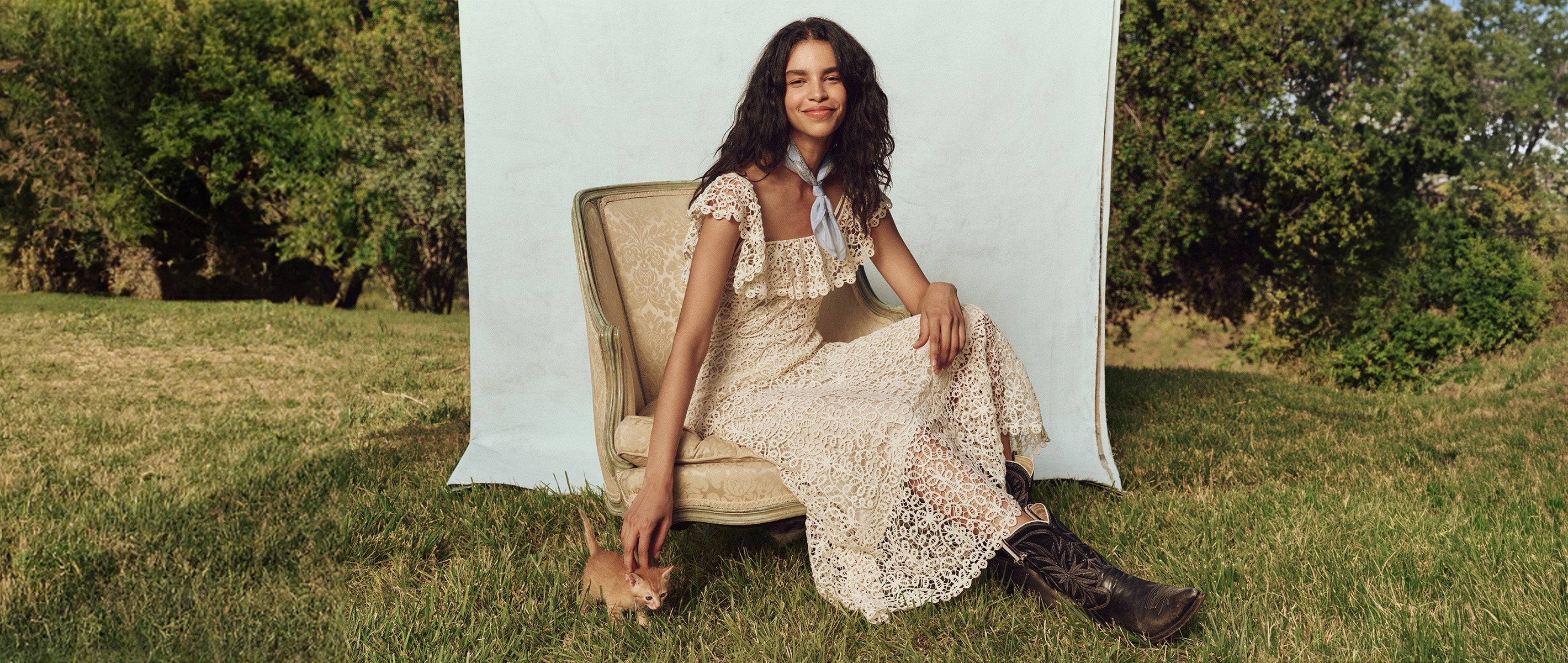 A person in a lace dress sits on a chair outdoors, petting a small dog on the grass, with trees and blue sky in the background.