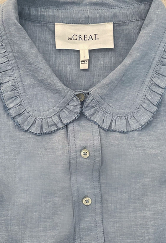 Close-up of a light blue blouse with a ruffled collar and buttons down the front. The label reads "de.CREAT.