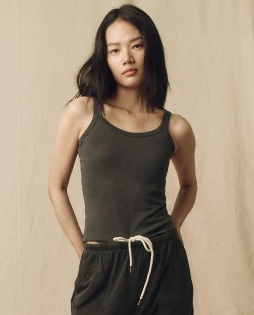 A person with long hair is wearing a dark tank top and pants with a drawstring, standing against a plain background.