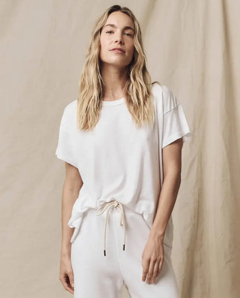 A person with long hair wearing a white t-shirt and pants stands against a beige backdrop.