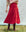 A person in a heart-patterned sweater, The Rhythm Skirt in Red Rose by THE GREAT. PS25, and brown boots stands gracefully on grass with a white backdrop, exuding a chic sense of fashion.