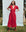 A person poses outdoors in THE GREAT. PS25's The Veranda Dress - Red Rose, stylishly paired with brown boots, standing on grass against a light blue backdrop.