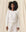 A person with curly hair stands against a beige backdrop, wearing a white long-sleeve top that looks like The Shrunken Sweatshirt from THE GREAT. FALL 24 KNITS in True White, paired with matching drawstring pants.