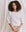 A person with curly hair, wearing 'The Shrunken Sweatshirt' – Washed White by THE GREAT. CORE KNITS, stands in front of a white fabric background.