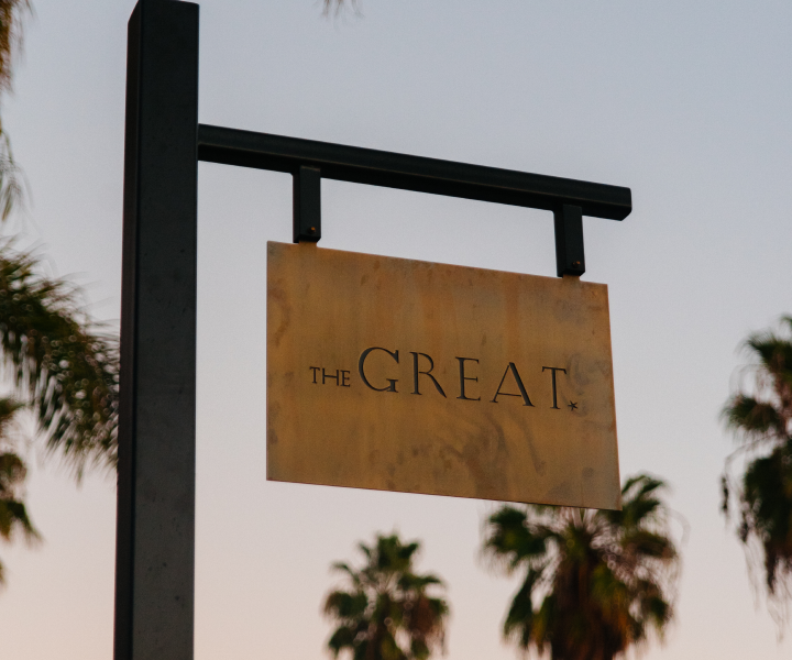 A sign reading "THE GREAT" hangs on a metal frame, with palm trees and a twilight sky in the background.