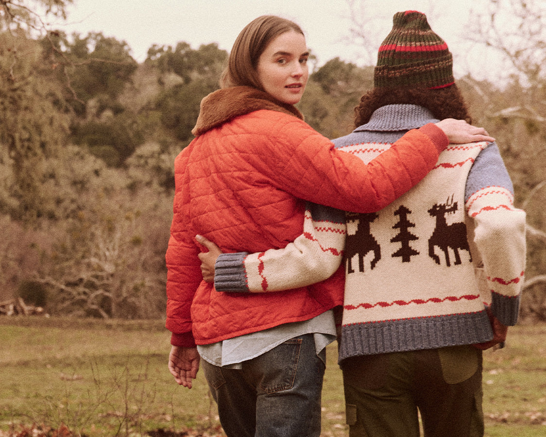 Two people in warm clothing walk arm in arm outdoors, one in a red jacket and the other in a patterned sweater with reindeer and trees. .